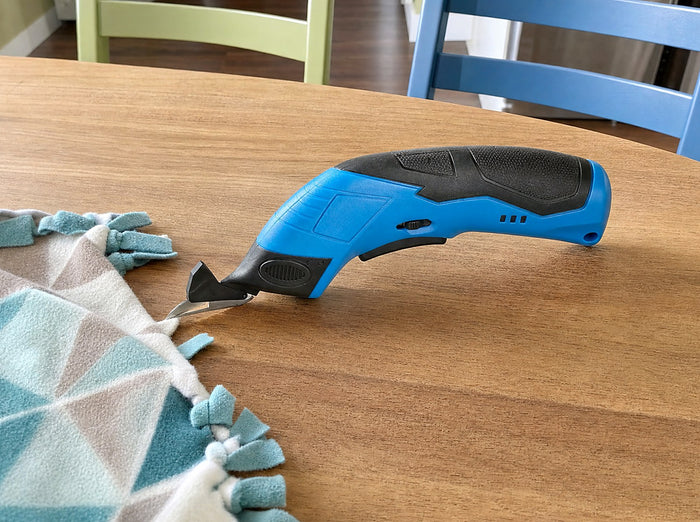 Exact blue shears alone beside a half-knotted fleece blanket edge on a breakfast table, two child-sized craft chairs empty in background; casual phone shot from high left in clear late-morning kitchen light, product new and fully visible