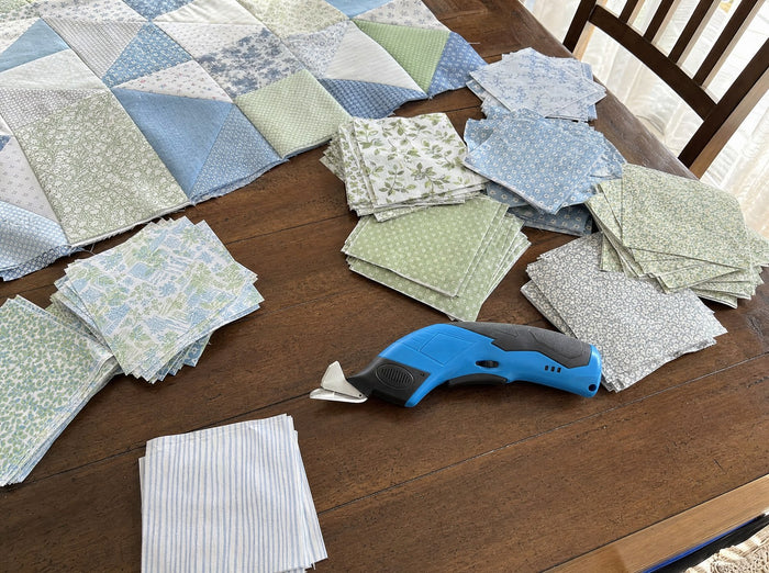 Exact blue shears alone on a folded quilt top at a clean dining table with many cut cotton squares, casual phone photograph from low front-right, bright cloudy afternoon light through patio glass; no styled arrangement